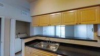 This single-floor space includes a well-appointed kitchen that serves as a central functional area. The kitchen features warm maple-toned cabinetry with raised panel doors and brushed metal hardware, offering generous upper storage. Dark granite-style countertops provide a strong contrast against the lighter cabinetry, creating a classic, practical aesthetic. A stainless steel double-basin sink is positioned along the counter with a standard faucet setup, while a reflective backsplash adds a subtle brightness to the workspace, bouncing light from a nearby window. Overhead ventilation is integrated into the ceiling, and electrical outlets are conveniently distributed along the counter. Adjacent to the kitchen, a dedicated laundry area is partially visible, suggesting thoughtful utility planning within the layout. The overall design is straightforward and functional, with neutral wall tones and consistent cabinetry throughout lending the space a cohesive, understated character. The single-floor layout ensures easy accessibility and efficient use of the available footprint.