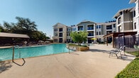 This two-floor residential community is defined by its impressive outdoor amenity space, centered around a generously sized resort-style swimming pool with a distinctive freeform shape. The pool area is a standout feature, complete with in-water lounge chairs that invite relaxation, a separate shallow spa zone, and stone-accented water features that add a refined, natural aesthetic. The surrounding pool deck is expansive, offering ample space for sunbathing with a collection of lounge chairs and bright yellow patio umbrellas positioned throughout. Wooden pergolas provide shaded retreats along the perimeter, complemented by a covered pavilion structure ideal for outdoor gatherings. The courtyard is enclosed by decorative iron fencing with gated access points, ensuring privacy and security. Mature trees and carefully maintained greenery, including trimmed hedges and a living green wall, add lush visual interest to the hardscaped areas. A cozy fire pit seating area is tucked into a landscaped nook near the building entrance, offering an intimate outdoor lounge space. The buildings themselves feature a combination of board-and-batten siding, limestone-clad bases, and private balconies that overlook the courtyard below. The overall ambiance is that of a well-appointed, amenity-rich community designed for outdoor living.