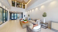 Located at 451 Hialeah Dr in Hialeah, Florida, the Shoma Village leasing office is a single-story facility designed to make a lasting first impression. The entrance welcomes visitors through floor-to-ceiling glass double doors framed in deep navy, flooding the interior with abundant natural light and offering clear views of the surrounding landscape.

Inside, the reception area is anchored by a sleek, modern desk set against a striking chevron-patterned wood accent wall bearing the Shoma Village branding. Warm amber pendant lights hang above, adding an inviting, boutique-style ambiance. Adjacent private offices with glass-panel walls and contemporary geometric light fixtures provide functional meeting spaces while maintaining an open, transparent feel.

The main lounge area commands attention with soaring double-height ceilings accented by exposed wood beams and a statement chandelier. Large clerestory windows flood the space with natural light, enhancing the airy, voluminous atmosphere. Light-toned large-format tile flooring runs throughout, complementing the crisp white textured walls. Comfortable neutral seating arrangements, accented with blue and orange throw pillows and tropical greenery, create a welcoming, resort-inspired setting. The overall design reflects a clean, contemporary coastal aesthetic, blending sophisticated architectural details with warm, approachable furnishings throughout.