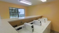 This single-floor shared laundry facility is designed with practicality and convenience in mind. The room features warm yellow-painted walls that create a welcoming atmosphere, complemented by bright overhead fluorescent lighting that ensures excellent visibility throughout the space. A row of modern front-loading washing machines lines the wall, providing ample laundry capacity for multiple users. Adjacent to the machines, a long, light-colored laminate folding counter runs the length of the room, offering generous workspace for sorting and folding laundry. Electrical outlets are conveniently positioned along the wall for added utility. A large multi-pane window floods the room with natural light and provides a pleasant view of the lush greenery and neighboring buildings outside, enhancing the overall ambiance of the space. The layout is thoughtfully organized to allow easy movement between the machines and the folding area, making laundry tasks efficient and straightforward. The clean, minimalist design and functional arrangement make this laundry room a practical and comfortable shared amenity.