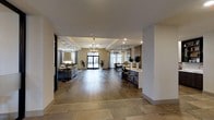 This single-floor space offers an open-concept layout that effortlessly connects multiple functional areas, creating a sense of expansiveness and flow. The entryway transitions from large-format stone tile flooring into warm, wide-plank hardwood that extends throughout the main living areas, grounding the space in natural texture and warmth.

The lounge area, visible to the left, features soft seating arrangements and a large floor lamp, complemented by floor-to-ceiling drapes and a reflective wall that amplifies the sense of space. An elegant orb chandelier serves as a focal point overhead, adding a touch of refined character to the room.

Toward the center, a set of glass-paned double doors floods the interior with natural light, framed by sheer curtains that soften the brightness. Just beyond, a dining area is illuminated by pendant lighting suspended from above, offering an inviting setting for gathering.

To the right, a kitchen counter with bar-height seating and pendant lights above defines the culinary zone, finished with clean cabinetry and a light countertop surface. Recessed lighting throughout ensures even, ambient illumination across the entire space.

The overall ambiance is modern yet welcoming, with a neutral color palette, thoughtful lighting design, and cohesive finishes that lend the space a polished and functional character.