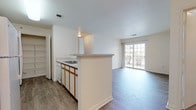 This single-floor unit offers a practical and comfortable layout designed for easy living. The open-plan kitchen and living area form the heart of the space, with the kitchen featuring white cabinetry accented by warm wood-tone trim, a smooth-top electric range, a built-in dishwasher, a stainless steel sink, and light-toned countertops. A peninsula with a breakfast bar separates the kitchen from the living area, while a dedicated walk-in pantry provides generous storage. The living area benefits from natural light streaming through a large sliding glass door that opens to the outdoors, and the space is finished with durable wood-style flooring throughout. The bedrooms are generously sized, carpeted for comfort, and include ample closet space. One bedroom features a walk-in closet, adding practical storage. The bathroom is clean and functional, offering a full soaking tub and shower combination with chrome fixtures, a wood-trimmed vanity with a mirror, and wood-style flooring. Throughout the unit, neutral white walls and consistent finishes create a cohesive, move-in-ready aesthetic. Ceiling ventilation and recessed lighting are present throughout, contributing to an overall sense of brightness and practicality.