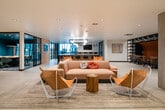 This thoughtfully designed two-floor amenity space combines contemporary architecture with a warm, layered aesthetic. On the main level, an expansive open-plan lounge serves as the social hub, furnished with plush blush sofas, leather sling chairs, and rustic wood side tables atop textured area rugs. Teal accent walls and globe pendant lighting add vibrant pops of color and warmth throughout. A generously sized kitchen and bar area anchors one end of the floor, featuring large dark-topped islands with built-in seating, globe and cage-style pendant lights, and large windows that draw in natural light. Adjacent to the lounge, a glass-enclosed conference room with black steel framing provides a sleek, private meeting environment visible from the common areas. A built-in banquette with orange upholstery and sculptural pendant fixtures lines the corridor leading toward the entrance. The second floor offers a quieter retreat, with warm wood-paneled walls, patterned carpet, and intimate seating alcoves divided by partial partitions — ideal for focused work or relaxed conversation. Dramatic floor-to-ceiling glazing on the upper level overlooks a striking multi-story atrium filled with hanging pendant clusters, connecting the two floors visually and reinforcing the space's open, airy character.