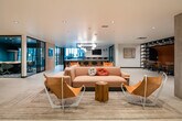 This thoughtfully designed two-floor amenity space combines contemporary architecture with a warm, layered aesthetic. On the main level, an expansive open-plan lounge serves as the social hub, furnished with plush blush sofas, leather sling chairs, and rustic wood side tables atop textured area rugs. Teal accent walls and globe pendant lighting add vibrant pops of color and warmth throughout. A generously sized kitchen and bar area anchors one end of the floor, featuring large dark-topped islands with built-in seating, globe and cage-style pendant lights, and large windows that draw in natural light. Adjacent to the lounge, a glass-enclosed conference room with black steel framing provides a sleek, private meeting environment visible from the common areas. A built-in banquette with orange upholstery and sculptural pendant fixtures lines the corridor leading toward the entrance. The second floor offers a quieter retreat, with warm wood-paneled walls, patterned carpet, and intimate seating alcoves divided by partial partitions — ideal for focused work or relaxed conversation. Dramatic floor-to-ceiling glazing on the upper level overlooks a striking multi-story atrium filled with hanging pendant clusters, connecting the two floors visually and reinforcing the space's open, airy character.
