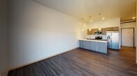 This contemporary single-floor unit offers a thoughtfully designed open-concept layout that seamlessly connects the kitchen, dining, and living areas. The kitchen is a standout feature, equipped with stainless steel appliances including a range, microwave, and refrigerator, complemented by dark wood-grain cabinetry, a bright white quartz countertop island, a double undermount sink, and stylish pendant lighting that hangs from soaring ceilings. The open living area extends toward a sliding glass door that leads to a private balcony, filling the space with natural light and outdoor views. Dark hardwood-style flooring runs throughout the main living areas, adding warmth and a modern aesthetic. Exposed ductwork along the upper walls and ceiling lends an industrial design element that adds character to the space. The bedroom features plush carpeting, a window with natural light, and exposed ductwork continuing the industrial theme. The bathroom is clean and functional, offering a full soaking tub and shower combination with brushed nickel fixtures. A dedicated in-unit laundry area adds everyday convenience. Overall, the unit presents a cohesive modern-industrial aesthetic with high ceilings, quality finishes, and a practical, open flow throughout.