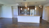 This single-floor space showcases a well-designed open-concept layout, with the kitchen serving as the central focal point. The kitchen features a large island with light granite countertops, providing generous workspace and a natural gathering area. Rich, dark hardwood flooring extends throughout the space, creating a warm and cohesive aesthetic.

The kitchen is outfitted with stainless steel appliances, including a French door refrigerator, gas range, and built-in microwave. Dark walnut-toned cabinetry lines the walls, offering substantial storage while adding depth and contrast against the lighter countertops and walls. Three pendant lights hang above the island, complementing the flush-mount ceiling fixtures to provide layered, functional lighting throughout the kitchen.

The open layout connects the kitchen seamlessly to the adjacent living areas, allowing for a fluid flow between spaces. Doorways visible from the kitchen suggest additional rooms beyond, with light-colored walls and clean lines maintaining a consistent, modern aesthetic throughout. The combination of hardwood floors, granite surfaces, and quality finishes gives the space a polished, contemporary character.