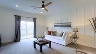 This single-story home showcases a thoughtfully designed living room that blends modern farmhouse aesthetics with functional comfort. The space is anchored by a striking white board-and-batten accent wall, which adds architectural depth and texture while serving as a backdrop for a rustic, horizontal wall art piece that enhances the room's character.

A plush white sofa dressed with earth-toned and patterned throw pillows sits at the center of the room, complemented by a solid dark wood coffee table that grounds the space. Overhead, a stylish ceiling fan with natural wood-toned blades and a matte black finish provides both visual interest and practical airflow.

The living area benefits from excellent natural light, courtesy of large sliding glass doors dressed with soft grey drape panels. The doors open to an outdoor area, blurring the line between interior and exterior living. Neutral carpet flooring and crisp white walls create a clean, airy ambiance throughout. Recessed lighting supplements the natural light, ensuring the space remains bright and welcoming at any hour.