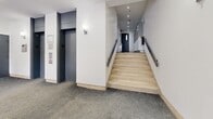 Located at 3455 Rue Aylmer in the heart of Montréal's vibrant Milton-Parc neighbourhood, this ground-floor common area serves as the primary entry and circulation hub of the building. The space is composed of a welcoming lobby and a connecting hallway, designed with clean, modern finishes throughout.

The entrance lobby greets residents and visitors with large glass doors that flood the interior with natural light and offer direct views onto Rue Aylmer. The flooring transitions from light-toned marble-style tiles near the entrance to durable grey carpet in the main circulation areas. White walls and recessed ceiling lighting contribute to a bright, tidy atmosphere. A small table and chair are positioned near the entry vestibule, providing a practical waiting or package-receiving area.

Leading from the lobby, the hallway provides access to the building's key vertical circulation elements: a compact elevator with stainless steel interior finishes and a staircase featuring light stone steps and sleek metal handrails. Wall-mounted sconces add warm accent lighting along the corridor.

At the far end of the hallway, a dedicated mail and parcel area includes Canada Post parcel lockers and a secure intercom panel, alongside unit access doors marked 102–103. A fire alarm station and security camera are also present, reflecting attentive safety standards throughout the common space.