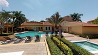 This single-story property showcases a well-appointed outdoor amenity area centered around a large, rectangular swimming pool with clear blue water. The surrounding pool deck is finished in light-colored pavers and furnished with multiple lounge chairs and reclining sun chairs, providing ample space for relaxation. Neatly trimmed hedgerows and tropical plantings, including a prominent palm tree, border the pool area, adding a lush, resort-like atmosphere.

The adjacent clubhouse structure features a distinctive Mediterranean-inspired design, highlighted by a sloped terracotta tile roof and warm stucco exterior walls finished in a soft tan hue. Large windows face the pool, allowing natural light into the interior while maintaining visual connectivity between indoor and outdoor spaces. A secondary structure to the right of the pool appears to house restroom or utility facilities, also finished in matching stucco and tile roofing to maintain architectural consistency.

The overall ambiance is bright and inviting, with an open-air layout that takes full advantage of the sunny climate. Mature trees in the background provide a natural privacy screen, enhancing the sense of a secluded retreat. The cohesive design and well-maintained landscaping give this outdoor space a polished, community-oriented character.