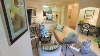 This single-floor residence showcases a thoughtfully designed open-concept layout that brings together the living, dining, and kitchen areas in one cohesive and functional space. The living area is comfortably appointed with a neutral-toned sofa, accent armchairs, and a round coffee table adorned with decorative blue vases, creating a welcoming focal point. Warm, light-toned walls and plush carpeting contribute to the space's inviting ambiance throughout. A dining nook with bar-style seating sits adjacent to the living area, making it ideal for casual meals or entertaining. The kitchen features dark cabinetry contrasted against light countertops and is equipped with stainless steel appliances, including a refrigerator. A pendant track lighting fixture illuminates the kitchen area, adding a modern touch. Crown molding details lend a refined architectural character to the space. A hallway visible from the main living area leads to additional rooms, hinting at the practical flow of the floor plan. The overall design balances comfort and style, with coordinated furnishings in blue, green, and neutral tones that create a harmonious and livable environment throughout.