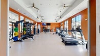 Located at 100 Conference Centre Dr in Cartersville, Georgia, this single-floor fitness center offers a generously sized, thoughtfully designed exercise facility. The space is divided into two distinct zones connected by a wide interior passage: a bright, open stretch and functional training area, and a larger main gym floor.

The stretch room features light-textured walls, smooth flooring, a ceiling fan, and a glass door with transom windows that open to an outdoor pool area, allowing natural light to pour in. Plyo boxes, resistance bands, yoga mats, and a color-coded dumbbell rack make it ideal for warm-ups and flexibility work.

The main gym area is defined by bold orange accent walls, elevated tray ceilings with multiple ceiling fans and pendant lighting, and floor-to-ceiling windows offering outdoor views. It is comprehensively equipped with a full row of treadmills and ellipticals, stationary and spin bikes, rowing machines, multi-use cable and strength machines, a punching bag, a tiered dumbbell rack, and medicine ball storage. Wall-mounted televisions and mirrored panels enhance the environment.

Barn-style interior doors connect sub-areas, and recessed lighting throughout ensures even illumination. The overall design is modern, spacious, and functional, creating a professional-grade workout environment.