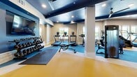 This modern fitness center offers a comprehensive workout facility thoughtfully organized into distinct training zones across a single, generously sized floor. The space features a striking design with bold navy blue ceiling accents contrasted against warm wood paneling and crisp white walls, creating an energetic yet polished atmosphere. Recessed lighting and ceiling fans ensure the space remains bright and comfortable throughout.

The cardio zone is lined with large windows that invite abundant natural light and outdoor views, housing a variety of equipment including treadmills, ellipticals, a stair climber, and a rowing machine. Full-length mirrors run along the walls, enhancing the sense of space and allowing for proper form checks during workouts.

The strength training area features a robust selection of equipment, including cable machines, Smith machines, free-weight racks, and a well-stocked dumbbell rack. A dedicated functional training zone offers TRX suspension systems, kettlebells, medicine balls, and resistance tools, all neatly organized on wall-mounted shelving.

Durable, color-zoned rubber flooring in yellow and blue delineates each training area while providing safety and comfort underfoot. Structural columns are neatly integrated into the layout, and glass-panel partitions add an architectural touch. The overall ambiance is professional and motivating, making this fitness center a fully equipped destination for a wide range of training needs.