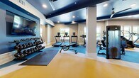 This modern fitness center offers a comprehensive workout facility thoughtfully organized into distinct training zones across a single, generously sized floor. The space features a striking design with bold navy blue ceiling accents contrasted against warm wood paneling and crisp white walls, creating an energetic yet polished atmosphere. Recessed lighting and ceiling fans ensure the space remains bright and comfortable throughout.

The cardio zone is lined with large windows that invite abundant natural light and outdoor views, housing a variety of equipment including treadmills, ellipticals, a stair climber, and a rowing machine. Full-length mirrors run along the walls, enhancing the sense of space and allowing for proper form checks during workouts.

The strength training area features a robust selection of equipment, including cable machines, Smith machines, free-weight racks, and a well-stocked dumbbell rack. A dedicated functional training zone offers TRX suspension systems, kettlebells, medicine balls, and resistance tools, all neatly organized on wall-mounted shelving.

Durable, color-zoned rubber flooring in yellow and blue delineates each training area while providing safety and comfort underfoot. Structural columns are neatly integrated into the layout, and glass-panel partitions add an architectural touch. The overall ambiance is professional and motivating, making this fitness center a fully equipped destination for a wide range of training needs.