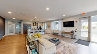 This single-floor communal space is designed with an open-concept layout that seamlessly connects multiple distinct activity zones. Light wood-tone flooring runs throughout, creating a cohesive and welcoming atmosphere across the entire space.

The kitchen area serves as a central hub, featuring an expansive granite-topped island with teal upholstered bar stools. Dark wood cabinetry pairs with stainless steel appliances, including a wall oven, microwave, refrigerator, and coffee station. Oversized pendant lights with metallic drum shades hang above the island, while decorative moss wall panels add a natural, organic accent.

The living area offers generous seating with a neutral sectional sofa accented by yellow throw pillows, arranged around a round coffee table atop a plush area rug. A wall-mounted flat-screen TV is positioned opposite the seating. A bold geometric textured accent wall in slate gray provides a striking architectural focal point nearby.

The game zone features a full-size shuffleboard table and a pool table, set against teal-painted walls. Multiple large windows dressed with sheer grommet curtains allow abundant natural light and outdoor views into this area.

Near the entrance, a tufted bench and console table create a welcoming transition point, with an adjacent wall-mounted TV and views to an outdoor patio beyond the glass door.