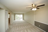 This single-floor residence offers a practical and well-organized layout across several distinct living areas. The main living space is generously proportioned, featuring neutral-toned walls with a contrasting gray accent panel, soft wall-to-wall carpeting, and a ceiling fan with integrated lighting. Sliding glass doors along one wall invite natural light and provide access to an outdoor balcony area, enhancing the sense of openness.

Adjacent to the living area, the kitchen is designed for functionality, featuring a peninsula-style breakfast bar that separates it from the carpeted living space. White upper cabinetry offers ample storage, while the kitchen is equipped with a black refrigerator, white electric range, and a dishwasher. Wood-look flooring runs throughout the kitchen, providing a clean contrast to the surrounding carpeted areas.

The bedroom is a well-sized, carpeted room with its own ceiling fan and a window that allows natural light to filter in. The bathroom includes a full-size soaking tub with a tiled surround, a curved shower rod, a dark-cabinetry vanity with a white countertop, a mirror, and wood-look flooring. A hallway connects these spaces, featuring a floor-level air vent and access to the bathroom through a framed doorway.