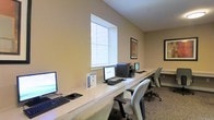 This single-floor business center offers a clean, functional workspace designed to accommodate multiple users simultaneously. A long, continuous work surface runs along the wall, providing generous desk space for several computer workstations complete with monitors, keyboards, and mice. A printer is conveniently stationed along the same counter, ensuring easy access for all users. Ergonomic rolling chairs accompany each workstation, promoting comfort during extended use. Natural light filters into the room through a large window fitted with horizontal blinds, creating a bright and pleasant working environment. The walls are finished in a warm neutral tone, contributing to a calm and focused atmosphere. Framed artwork adorns the walls, adding a subtle decorative touch to the otherwise professional setting. Carpeted flooring runs throughout the space, softening the room's acoustics and adding to its understated comfort. Overall, the business center is thoughtfully laid out to provide a productive and welcoming environment for anyone in need of a reliable workspace.