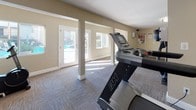 This single-floor fitness center is thoughtfully designed to provide a functional and comfortable workout environment. The space is finished with durable patterned carpet tiles throughout, complemented by warm taupe-toned walls and crisp white baseboards and column trim that lend a clean, polished appearance.

The workout area is generously sized and organized into distinct exercise zones. Cardio equipment, including treadmills and elliptical trainers, is positioned to take advantage of the natural light that streams in through multiple large windows and glass-panel doors. A full-length mirrored wall along one side of the room visually expands the space and aids exercisers in monitoring their form. Additional strength and resistance training equipment, including a multi-station cable machine and a weight bench, round out the facility's offerings.

Structural columns are integrated throughout the layout, adding architectural character while subtly defining different workout areas. Decorative touches such as tall floor planters with tropical greenery add warmth and a welcoming ambiance. Flush-mounted ceiling fixtures provide consistent overhead lighting throughout.

French doors along the perimeter open directly to an outdoor pool area, creating a seamless connection between the interior fitness space and the exterior amenities, while also allowing fresh air and ample sunlight to fill the room during the day.