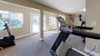 This single-floor fitness center is thoughtfully designed to provide a functional and comfortable workout environment. The space is finished with durable patterned carpet tiles throughout, complemented by warm taupe-toned walls and crisp white baseboards and column trim that lend a clean, polished appearance.

The workout area is generously sized and organized into distinct exercise zones. Cardio equipment, including treadmills and elliptical trainers, is positioned to take advantage of the natural light that streams in through multiple large windows and glass-panel doors. A full-length mirrored wall along one side of the room visually expands the space and aids exercisers in monitoring their form. Additional strength and resistance training equipment, including a multi-station cable machine and a weight bench, round out the facility's offerings.

Structural columns are integrated throughout the layout, adding architectural character while subtly defining different workout areas. Decorative touches such as tall floor planters with tropical greenery add warmth and a welcoming ambiance. Flush-mounted ceiling fixtures provide consistent overhead lighting throughout.

French doors along the perimeter open directly to an outdoor pool area, creating a seamless connection between the interior fitness space and the exterior amenities, while also allowing fresh air and ample sunlight to fill the room during the day.