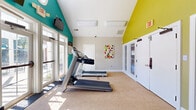 Located at 5613 Crenshaw Rd in Richmond, VA, this fitness center is a well-appointed, single-room exercise facility designed to support a well-rounded workout routine. The space features vaulted ceilings that lend an open, airy feel, while bold teal and yellow-green accent walls add an energizing visual character against the neutral gray and beige tones throughout.

Natural light flows generously into the room through large windows and a set of glass-paneled French doors, creating a bright and welcoming environment. The carpeted floor provides a comfortable surface underfoot, and the layout efficiently accommodates a variety of equipment including multiple treadmills, an elliptical trainer, a stationary bike, and a multi-station strength training machine — offering both cardio and resistance training options.

A wall-mounted flat-screen television is positioned for easy viewing during workouts, while a colorful abstract artwork on the wall adds a creative touch to the space. Overhead lighting fixtures supplement the natural light, ensuring consistent illumination throughout the day. Security is supported by a ceiling-mounted surveillance camera. The fitness center is accessible via multiple entry points, including the French doors that lead to an adjacent outdoor area, making this a functional and inviting space for daily exercise.