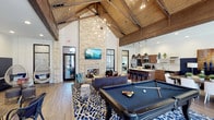 This impressive clubhouse is a single-floor gathering space designed to blend rustic charm with modern amenities. The centerpiece of the main lounge is a dramatic vaulted ceiling adorned with rich wood planking and exposed beam trusses, soaring to remarkable heights and anchored by a striking sputnik chandelier that fills the space with warm, ambient light. A floor-to-ceiling stacked stone fireplace serves as a bold focal point, complemented by a wall-mounted television above the mantle — creating a cozy yet grand atmosphere. The lounge area features stylish, eclectic furnishings, including a hanging chair, plush seating, and a bold patterned area rug that adds a pop of personality to the hardwood floors. A full-size billiard table takes center stage, making the space ideal for social gatherings and recreation. Adjacent to the lounge, a fully equipped kitchen and bar area offers ample counter space, modern appliances, a large island with bar seating, and statement pendant lighting — perfect for hosting events. Large black-framed windows throughout allow natural light to pour in, connecting the interior with the outdoors. Overall, this clubhouse delivers a welcoming, upscale environment built for community and entertainment.