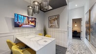 Located at 622 Brigham Rd in Greensboro, NC, this thoughtfully designed single-floor space functions as a sophisticated communal and transitional area. The bold black-and-white geometric tile flooring establishes a strong visual foundation throughout, creating a cohesive flow from the welcoming entry to the interior corridors. The entry features glass-paneled double doors that invite natural light and views of the exterior, complemented by a sleek charcoal bench and large-scale contemporary artwork on the walls. Moving inward, the hallway connects multiple private rooms and workspaces, each accessible through crisp white-framed doorways. A built-in desk area provides a convenient workstation niche along the corridor. The central meeting area is a standout feature, anchored by a white conference table surrounded by yellow accent chairs and dramatically lit by a trio of sculptural wire cage pendant lights suspended from a dark ceiling cutout. Wainscoting lines the walls, adding architectural refinement. A lounge nook offers additional seating beneath a grid of distinctive dark sculptural wall art, with natural light streaming in from a nearby window. The overall ambiance balances contemporary design with warm, curated details, making the space both functional and visually engaging.