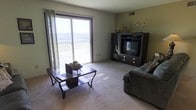 This single-floor space presents a welcoming and functional layout centered around a comfortable living area. The living room is a standout feature, bathed in natural light streaming through a large sliding glass door dressed with sheer curtains. The door opens to a balcony or patio area, framing a broad, scenic view of the surrounding landscape — including what appears to be an open waterfront or expansive outdoor scenery in the distance, lending the room an airy, open feel.

The living room is finished with neutral-toned carpeting that adds warmth underfoot, while the walls are painted in a soft, muted green that complements the overall calm ambiance. A dark wood coffee table anchors the seating area, accompanied by a plush gray sofa, creating a cozy and inviting atmosphere. An entertainment center in a matching dark wood finish houses a television and provides storage, serving as a functional focal point on the opposite wall.

Subtle decorative touches — including candelabra-style accents and small decorative pieces atop the entertainment unit — add character to the space. A ceiling vent indicates a central HVAC system for year-round comfort. Overall, the space combines practicality with a relaxed aesthetic, highlighted by its impressive natural light and scenic outdoor views.