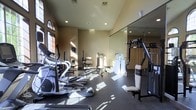 This fitness center offers a comprehensive workout environment designed to accommodate a range of exercise routines. The space is outfitted with a selection of cardio equipment, including elliptical machines prominently positioned near large windows that allow abundant natural light to fill the room. Strength training is well-supported by a multi-function cable machine and a dedicated free weights and bench press area. A full-length mirrored wall runs along one side, enhancing the sense of openness while allowing users to monitor their form during workouts. The flooring features durable, dark rubberized material suited for high-impact activity. Recessed lighting in the ceiling supplements the natural light from the tall arched windows, creating a bright and energizing atmosphere throughout the day. The warm neutral-toned walls and architectural details, including raised ceiling sections, contribute to a polished and welcoming environment. A water station is conveniently positioned within the space, ensuring easy access to hydration. Overall, the fitness center is thoughtfully arranged to maximize usability while maintaining an open, comfortable feel for all fitness levels.