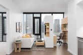 This modern urban residence showcases a thoughtfully designed single-floor layout with a clean, contemporary aesthetic throughout. The open-plan living area features rich hardwood flooring and floor-to-ceiling windows framed in dark metal, offering sweeping city skyline views that flood the space with natural light. The living room comfortably accommodates a sectional sofa and media unit, creating an inviting yet streamlined atmosphere.

The kitchen is well-appointed with flat-front cabinetry in a soft grey finish, dark stone countertops, and a distinctive geometric-patterned tile backsplash. A full suite of stainless steel appliances includes a gas range, built-in microwave, dishwasher, and a large French door refrigerator, catering to both everyday cooking and entertaining.

The bedroom benefits from corner-positioned floor-to-ceiling windows, providing panoramic city views and generous natural light, with plush carpeting underfoot for comfort. The bathroom features a full soaking tub with an overhead shower, dramatic dark-tiled surround, and wood-look tile flooring, creating a bold contrast against the white walls. Recessed lighting throughout contributes to a polished, cohesive ambiance across the entire space.