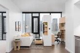 This modern urban residence showcases a thoughtfully designed single-floor layout with a clean, contemporary aesthetic throughout. The open-plan living area features rich hardwood flooring and floor-to-ceiling windows framed in dark metal, offering sweeping city skyline views that flood the space with natural light. The living room comfortably accommodates a sectional sofa and media unit, creating an inviting yet streamlined atmosphere.

The kitchen is well-appointed with flat-front cabinetry in a soft grey finish, dark stone countertops, and a distinctive geometric-patterned tile backsplash. A full suite of stainless steel appliances includes a gas range, built-in microwave, dishwasher, and a large French door refrigerator, catering to both everyday cooking and entertaining.

The bedroom benefits from corner-positioned floor-to-ceiling windows, providing panoramic city views and generous natural light, with plush carpeting underfoot for comfort. The bathroom features a full soaking tub with an overhead shower, dramatic dark-tiled surround, and wood-look tile flooring, creating a bold contrast against the white walls. Recessed lighting throughout contributes to a polished, cohesive ambiance across the entire space.