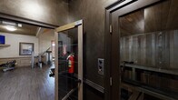 This fitness center in Madison, Alabama offers a comprehensive workout facility spread across a single floor, organized into distinct zones designed to accommodate a wide range of exercise needs. The main strength training area features soaring vaulted ceilings with warm wood accents and a coffered ceiling detail highlighted in rich amber tones, creating an inviting and upscale atmosphere. Dark hardwood-style flooring runs throughout this section, which is lined with an extensive selection of Life Fitness and other commercial-grade strength machines, free weights, cable systems, and a Smith machine. Full-length mirrors along the walls enhance the sense of openness and allow for form monitoring during workouts. A dedicated cardio room, finished with warm terracotta-toned walls, a natural wood-paneled vaulted ceiling, and light tile flooring, houses multiple treadmills, elliptical machines, stationary bikes, and stair climbers. Wall-mounted televisions are positioned for comfortable viewing during cardio sessions, and natural light filters in through windows with wood-slatted blinds. The facility also includes a glass-panel entryway corridor leading to additional workout areas, with safety equipment such as fire extinguishers readily accessible. Ceiling fans throughout contribute to a comfortable exercise environment.