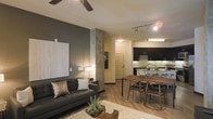 This thoughtfully designed single-floor space showcases a seamless open-concept layout that blends the living, dining, and kitchen areas into one cohesive, modern environment. The living area is anchored by a sleek dark sofa set against a bold charcoal accent wall, complemented by warm ambient lighting and a ceiling fan overhead that adds both function and style. A structural column with a textured concrete finish serves as a striking architectural focal point, adding an industrial edge to the contemporary aesthetic. The dining area features a warm-toned wooden table paired with mid-century modern chairs, creating an inviting space for gatherings. The kitchen is defined by dark cabinetry, under-cabinet lighting, and track lighting along the ceiling, giving it a clean and polished look. Light hardwood flooring flows consistently throughout the space, tying all areas together while enhancing the sense of openness and warmth. The neutral color palette, punctuated by contrasting dark tones and natural textures, creates a balanced and sophisticated ambiance. Subtle greenery adds a refreshing organic touch to the overall design, making this space both visually dynamic and welcoming.