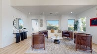 This thoughtfully designed single-floor professional office space blends modern aesthetics with functional layout. The main entrance greets visitors with soaring ceilings, floor-to-ceiling glass double doors, and sidelights that flood the entryway with abundant natural light, creating an immediately welcoming atmosphere.

The waiting lounge is a standout feature, furnished with rattan barrel chairs, a sculptural round coffee table, and a built-in window bench seat. Multiple large windows frame views of the surrounding greenery, enhancing the open and airy feel of the room. Light hardwood flooring flows consistently throughout the entire space, unifying each area with warmth and cohesion.

The front office area features dual reception desks with rich dark wood cabinetry, frosted-glass private office doors, and a striking blue abstract mural that serves as a bold visual focal point along the central corridor. This hallway leads deeper into the suite, where private offices provide quiet, focused workspaces.

A casual collaboration zone offers a bar-height table with wishbone-style seating beneath a woven rattan pendant light, accompanied by large windows and curated wall art — ideal for informal meetings or team gatherings. The break room features warm wood cabinetry, a white countertop, stainless steel appliances, and a dramatic blue abstract mural accent wall, adding an artistic energy to the everyday amenity space.