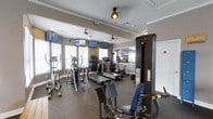 This fitness center is thoughtfully designed to support a comprehensive workout experience. The space is outfitted with a robust selection of cardio equipment, including multiple treadmills, elliptical trainers, and stationary bikes, all arranged to maximize usability and floor space. A prominent Cybex VR1 strength training machine anchors one side of the room, offering users a dedicated area for resistance-based exercise. The flooring features durable dark rubber matting, practical and resilient for high-traffic fitness use. Several wall-mounted flat-screen televisions are positioned throughout the room, allowing users to stay entertained or informed during their workouts. Ceiling fans provide continuous air circulation, contributing to a comfortable environment. Large windows fitted with blinds line the walls, inviting ample natural light while maintaining privacy and glare control. Crown molding along the upper walls adds a refined architectural touch that elevates the overall aesthetic of the space. A mirrored wall section reflects the room, enhancing the sense of openness. The warm, neutral wall tones create a clean and focused atmosphere well-suited for exercise. Overall, this fitness center combines functional equipment with thoughtful design to deliver a well-rounded and inviting workout environment.