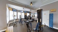 This fitness center is thoughtfully designed to support a comprehensive workout experience. The space is outfitted with a robust selection of cardio equipment, including multiple treadmills, elliptical trainers, and stationary bikes, all arranged to maximize usability and floor space. A prominent Cybex VR1 strength training machine anchors one side of the room, offering users a dedicated area for resistance-based exercise. The flooring features durable dark rubber matting, practical and resilient for high-traffic fitness use. Several wall-mounted flat-screen televisions are positioned throughout the room, allowing users to stay entertained or informed during their workouts. Ceiling fans provide continuous air circulation, contributing to a comfortable environment. Large windows fitted with blinds line the walls, inviting ample natural light while maintaining privacy and glare control. Crown molding along the upper walls adds a refined architectural touch that elevates the overall aesthetic of the space. A mirrored wall section reflects the room, enhancing the sense of openness. The warm, neutral wall tones create a clean and focused atmosphere well-suited for exercise. Overall, this fitness center combines functional equipment with thoughtful design to deliver a well-rounded and inviting workout environment.