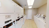 This single-floor communal laundry facility is designed with functionality and convenience in mind. The room is outfitted with an extensive lineup of commercial-grade washing machines and dryers, including stacked dryer units along one wall and a row of side-by-side washers and dryers lining the opposite side, providing ample capacity for multiple users simultaneously. Each machine features clearly labeled coin-operated or digital control panels, making operation straightforward and user-friendly. The space is finished with neutral beige walls and light-toned tile flooring, creating a clean and welcoming environment that is easy to maintain. Overhead, flush-mounted fluorescent light panels illuminate the room evenly, ensuring excellent visibility throughout. A solid interior door provides access to an adjoining utility or storage area, adding to the overall practicality of the space. The layout is thoughtfully organized, with sufficient aisle space between machine rows to allow comfortable movement and access. The overall ambiance is bright, tidy, and efficient, reflecting a well-maintained shared facility suited to meet the everyday laundry needs of its users.