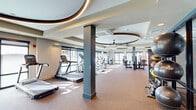 This well-appointed fitness center offers a generously sized, single-floor exercise space designed to accommodate a full range of workout routines. The room's architectural design is immediately striking, with distinctive circular recessed ceiling elements accented by integrated LED lighting strips that lend a modern, upscale aesthetic throughout the space. Large ceiling fans are centrally positioned within these circular ceiling features, ensuring effective air circulation across the floor.

Natural light floods the interior through expansive floor-to-ceiling windows along multiple walls, creating a bright and energizing atmosphere. The neutral-toned carpet flooring provides comfortable underfoot support across the entire exercise area.

The cardio section is well-equipped with multiple treadmills, elliptical trainers, stationary bikes, and a rowing machine, all arranged to maximize space and views to the outdoors. The strength training area features a variety of cable and resistance machines, free weights, benches, and a medicine ball rack, offering a comprehensive selection for users of all fitness levels. Full-length mirrors along one wall enhance the sense of openness while providing functional support for strength training. Structural columns are integrated cleanly into the layout, and the overall floor plan flows efficiently, ensuring ample room to move between equipment zones.