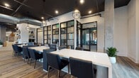 Located at 200 E Cama St in Charlotte, NC, this single-floor space masterfully combines industrial aesthetics with modern, curated design. The open layout is anchored by striking dark-painted ceilings with exposed ductwork and recessed lighting, contrasted by crisp white walls and wide-plank hardwood flooring throughout.

The lounge area is a standout, featuring an eclectic mix of seating including a teal velvet sofa, a bold crimson sectional, and neutral accent chairs arranged around a circular area rug. Tall, slender windows dressed in sheer white curtains flood the space with natural light, while a large sculptural wooden art piece serves as a dramatic focal point. An elegant tiered circular chandelier adds warmth and sophistication to the seating vignette.

The co-working zone is defined by long, sleek white work surfaces lined with charcoal upholstered chairs, all enclosed by floor-to-ceiling steel-framed glass partitions that maintain visual connectivity while providing a sense of structure. Globe pendant lights hang overhead, complementing the industrial-chic theme.

A separate enclosed room, visible through the glass walls, offers additional private workspace or meeting space. Together, these areas create a dynamic, multi-functional environment ideal for collaboration, focused work, and social gathering.