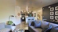 This single-story residence features an open-concept layout that connects the living area and kitchen, creating a cohesive and functional environment. The kitchen is outfitted with dark espresso cabinetry, black appliances, and a central island with bar-height seating, making it a practical and stylish focal point. A combination of pendant lighting and a track light fixture provides layered illumination throughout the space. The living area is comfortably appointed and flows naturally from the kitchen, promoting an inclusive, social atmosphere. Warm hardwood-style flooring extends throughout, tying the spaces together visually. Rustic decorative elements, including mounted animal head wall accents and circular wooden décor, add character and a lodge-inspired aesthetic. A hallway visible from the main living area leads to additional rooms, which appear to include built-in shelving and further living or sleeping spaces. The neutral wall palette, accented by a bold dark feature wall near the hallway entrance, balances the warmth of the décor with a contemporary edge. Overall, the space offers a welcoming and well-designed environment suited for both everyday living and entertaining.