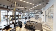 This expansive, single-floor fitness center is designed to accommodate a comprehensive range of workout disciplines within a sleek, contemporary setting. The space is defined by its bold geometric-patterned flooring in neutral tones of cream, taupe, and gray, which visually delineates the various exercise zones throughout the facility.

The cardio area features an extensive row of Precor treadmills, elliptical trainers, and stair climbers, each equipped with individual screens for entertainment. Large windows along the perimeter flood this section with abundant natural light and offer views of the surrounding greenery. The strength training zone is anchored by a series of heavy-duty power racks stocked with barbells, weight plates, and benches, alongside a full array of Precor selectorized cable and resistance machines suited for targeted muscle training.

A dedicated free weights area provides dumbbells, medicine balls, and functional training accessories such as battle ropes and TRX suspension systems. Glass-walled studio rooms at the perimeter offer separate spaces for group fitness, cycling, or stretching, fitted with mirrored walls and wood-tone flooring. Overhead, a dynamic arrangement of linear LED lighting crisscrosses the ceiling, creating an energetic atmosphere while ensuring bright, even illumination throughout the entire facility.