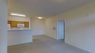 This single-floor unit offers an open-concept layout that seamlessly connects the kitchen and living area. The living space is finished with wall-to-wall neutral carpet, contributing to a quiet and comfortable atmosphere. Walls are painted in a soft, light tone throughout, enhancing the overall sense of brightness in the space.

The kitchen is positioned along one side of the open layout, separated by a raised breakfast bar that provides a natural division between cooking and living areas. It features warm-toned wood cabinetry, an overhead microwave, and under-cabinet lighting. The raised counter serves both as a functional workspace and an informal dining surface.

The main living area is generously sized, with smooth ceilings fitted with flush-mount lighting fixtures and a smoke detector. A wall-mounted HVAC vent is centrally located, ensuring efficient climate distribution throughout the space. A paneled interior door visible toward the rear of the unit leads to an adjoining room, suggesting additional private living spaces beyond. Electrical outlets and light switches are thoughtfully placed along the walls for convenience.