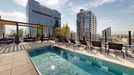 Located at 633 South LaSalle Street in Chicago's South Loop neighborhood, this rooftop terrace is an elevated outdoor amenity space offering a compelling blend of modern design and urban scenery. The terrace is finished with large-format light-toned pavers throughout, creating a clean, contemporary aesthetic that complements the surrounding architectural cladding of gray composite panels and expansive floor-to-ceiling glass curtain walls.

At the heart of the terrace sits a sleek, rectangular swimming pool with calm, clear water and clearly marked depth indicators, flanked by a generous sundeck lined with padded chaise lounges — ideal for relaxation while taking in the panoramic Chicago skyline. Lush greenery in large white cylindrical planters and raised planting beds adds softness and life to the space, creating a sense of an urban garden retreat.

A covered pergola structure with a wood-slat ceiling defines a shaded lounge area furnished with comfortable seating, offering a more intimate setting within the open terrace. Nearby, benches and bistro-style tables and chairs provide additional gathering spots. The terrace benefits from abundant natural light and unobstructed, sweeping views of the surrounding South Loop high-rises and the broader Chicago cityscape, making it a distinctive and functional outdoor destination.