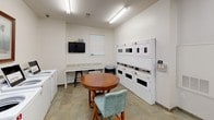 Located at 3920 Ivy Summit Ct in Cumming, Georgia, this single-floor shared laundry facility is designed to provide a clean, functional, and comfortable experience for its users. The space is organized into distinct zones that efficiently accommodate the full laundry process from start to finish.

The washing area features a row of top-loading commercial washers, each clearly numbered for easy identification, accompanied by a front-loading washer-dryer unit. Stacked commercial dryers line one wall, offering generous drying capacity. The machines are well-maintained and set against bright white walls, contributing to the space's clean and orderly appearance.

Generous granite-topped folding counters span multiple sections of the room, providing ample surface area for sorting and folding laundry. Wooden stools are positioned along the counters for added convenience. The flooring throughout is durable stone-look tile, practical for a high-use facility.

A wall-mounted flat-screen television adds a touch of comfort, making wait times more enjoyable. A round wooden table with seating offers a relaxed area to sit while laundry cycles run. Decorative framed artwork on the walls adds a welcoming touch to the otherwise utilitarian space. Overhead fluorescent lighting ensures the entire facility is well-lit, and a windowed door allows for natural light and easy access.