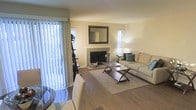 This single-floor residence centers around a welcoming living area designed for both comfort and functionality. The space features warm hardwood flooring throughout, lending a cohesive and polished look to the interior. A tile-surround fireplace serves as the room's focal point, topped with an ornate framed mirror that adds depth and character to the neutral, cream-toned walls.

Natural light fills the space generously, streaming in through multiple windows fitted with horizontal blinds and a large sliding glass door dressed with vertical blinds, which provides an easy transition to the outdoors. The living area comfortably accommodates a plush sofa accented with teal decorative pillows, positioned around a glass-top coffee table with a distinctive X-frame base resting on a neutral area rug.

A dining area is partially visible near the sliding glass door, hinting at a practical open layout that connects living and dining zones seamlessly. Soft ambient lighting from a table lamp complements the natural light, creating a warm and inviting atmosphere. The overall design is clean, understated, and move-in ready.