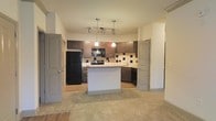 This single-floor space showcases an open-concept layout that seamlessly connects the kitchen and living area. The kitchen is a standout feature, equipped with rich dark wood cabinetry that provides ample storage, complemented by sleek black appliances including a full-size refrigerator, range, microwave, and dishwasher. A bold geometric tile backsplash adds a modern design accent behind the cooking area, while light-toned countertops provide a subtle contrast to the darker elements throughout.

A central kitchen island serves as a functional divider between the kitchen and the adjoining living space, offering additional prep space and a natural gathering point. Pendant lights hang above the island, contributing warm, focused illumination, while a track lighting fixture overhead ensures the kitchen remains well-lit throughout. The transition from the hard flooring in the kitchen to plush carpeting in the living area creates a clear and comfortable distinction between the two zones. Neutral wall tones and a light ceiling keep the space feeling bright and open. Two interior doors, finished in a soft grey, provide access to additional rooms within the unit.