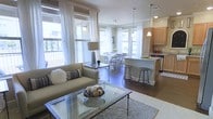 This single-floor residence features an open-concept layout that integrates the living, dining, and kitchen areas into one cohesive, flowing space. Large windows dressed with sheer white curtains line the walls, flooding the interior with abundant natural light and offering views of the surrounding neighborhood. The living area is anchored by a neutral-toned sofa accented with decorative pillows and paired with a glass-topped coffee table resting on a textured area rug, creating a comfortable and stylish focal point. Rich hardwood flooring extends throughout, adding warmth and continuity across the space. The dining area accommodates a white dining table with matching chairs, positioned near a window-lined corner that enhances the airy atmosphere. Transitioning seamlessly into the kitchen, a granite-topped breakfast bar with bar stools serves as both a functional workspace and casual seating area. The kitchen features light wood cabinetry, stainless steel appliances, and pendant lighting that adds a refined touch. The overall ambiance is bright, modern, and welcoming, with a cohesive neutral palette and thoughtful design details throughout.
