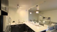 This single-floor residence offers a thoughtfully designed open-concept layout that seamlessly connects the kitchen and living areas. The kitchen is a standout feature, showcasing rich dark cabinetry that contrasts elegantly with bright white countertops. Stainless steel appliances, including a full-size refrigerator, microwave, and range, provide a sleek and functional cooking space. A double-basin sink is set into the expansive L-shaped counter, offering ample prep space. The kitchen island extends into a breakfast bar, accommodating two modern white bar stools — an ideal spot for casual dining or entertaining. Pendant lighting hangs above the bar, adding warmth and a contemporary touch to the space, while track lighting in the adjacent area provides flexible illumination. The open flow into the living room creates a spacious and airy atmosphere, enhanced by light wood-tone flooring that runs throughout. The neutral wall tones contribute to a clean, modern aesthetic. A hallway visible from the main living area suggests additional rooms beyond, completing the functional layout of this well-appointed single-level residence.