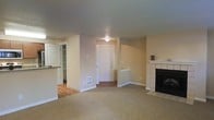 This single-floor residence offers a comfortable and functional layout with a clean, neutral aesthetic throughout. The living area is finished with wall-to-wall carpet and anchored by a classic tiled fireplace with a white mantel, creating a warm and inviting focal point. Neutral beige walls and flush ceiling lighting give the space a bright, understated feel. A paneled front door provides a defined entryway, seamlessly connecting to the main living space. Adjacent to the living area, hardwood flooring transitions into what appears to be a kitchen and pantry zone, where white cabinetry with open shelving offers practical storage. The open sightlines between the living and kitchen areas contribute to a sense of flow and connectivity throughout the home. A floor vent indicates a central HVAC system for year-round climate control. The overall design is straightforward and move-in ready, with a traditional character reflected in the paneled doors, tiled fireplace surround, and classic trim details. This space is well-suited for those seeking a low-maintenance, single-level home with functional living areas and timeless finishes.