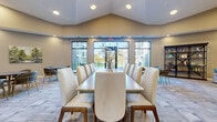 Located at 6620 Chestnut St in Lino Lakes, MN, this single-floor community space offers a thoughtfully designed open layout combining a fully equipped kitchen and expansive living area. The kitchen is outfitted with crisp white shaker cabinetry, stainless steel appliances including a full-size refrigerator, wall oven, and built-in microwave, complemented by stone-look countertops, a deep utility sink, and open shelving. Stylish gold-accented pendant lights hang above the kitchen island, which features upholstered bar seating in a contemporary blue-gray tone. The living area encompasses multiple functional zones, including a formal dining space anchored by a long conference-style table with cushioned high-back chairs beneath a vaulted ceiling, and a relaxed lounge area centered around a linear fireplace set into a striking stacked-stone accent wall with a mounted flat-screen television above. Patterned carpet tiles throughout the living zones add texture and warmth, while large windows in the dining area flood the space with natural light and offer views of the surrounding outdoor grounds. The overall ambiance is modern and welcoming, with a neutral color palette, quality finishes, and versatile seating arrangements that support both social gatherings and quiet relaxation.