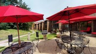 This inviting outdoor courtyard is set within a classic multi-building complex constructed of warm brick, featuring pitched rooflines and a well-maintained, landscaped environment. The centerpiece of the space is a generously sized concrete patio that serves as an al fresco gathering area, furnished with wrought iron tables and chairs arranged beneath a series of vibrant red market umbrellas that provide ample shade on sunny days. A gas grill station is conveniently positioned at the heart of the patio, making it ideal for outdoor cooking and entertaining. Large decorative planters with seasonal flowers add color and character to the hardscape. The patio transitions seamlessly to manicured green lawns that stretch between the surrounding buildings, with mature trees lining the perimeter and a winding pathway leading further into the grounds. The overall ambiance is relaxed and communal, designed to encourage outdoor socializing and leisure. The combination of durable, classic architecture, thoughtfully arranged seating, and lush green surroundings creates a welcoming and functional outdoor environment suited for both casual gatherings and quiet relaxation.