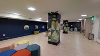 Located at 1530 Locust Street in the heart of Philadelphia, PA, this single-floor space is thoughtfully designed to engage and inspire young visitors. The open layout features light hardwood flooring and deep navy-blue accent walls that create a bold, inviting atmosphere throughout.

Among the most striking design elements are the large structural columns wrapped in vivid, nature-inspired mosaic artwork, depicting oversized leaves and geometric shapes in rich greens, yellows, and purples. These artistic installations serve as visual focal points that bring energy and creativity to the space.

The room is furnished with modular, colorful hexagonal seating in shades of orange, teal, gray, and yellow, allowing for flexible arrangement to suit a variety of activities. Circular mirrors are mounted along the navy walls, adding depth and a playful visual element to the room.

Toward the back of the space, shelving units and organizational furniture provide practical storage for materials and supplies, while a clock and additional activity areas suggest a structured yet dynamic environment. Recessed and flush-mounted ceiling lighting ensures the space is well-lit throughout. The overall ambiance is lively, imaginative, and purpose-built to support an engaging, child-centered experience.